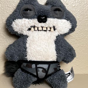 May include: A plush toy wolf with gray fur, white chest, and a black nose. The wolf has a wide grin with visible teeth and is wearing camouflage print underwear. The toy has a tag attached.