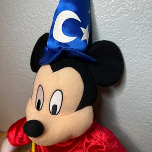 K&ouml;nnte beinhalten: Ein Mickey Mouse-Pl&uuml;schtier, das als Zauberer verkleidet ist. Das Spielzeug hat einen blauen Hut mit wei&szlig;en Sternen und einem Mond, eine rote Robe und einen gelben Seilg&uuml;rtel. Das Gesicht ist beige mit schwarzen Augen und einer schwarzen Nase.