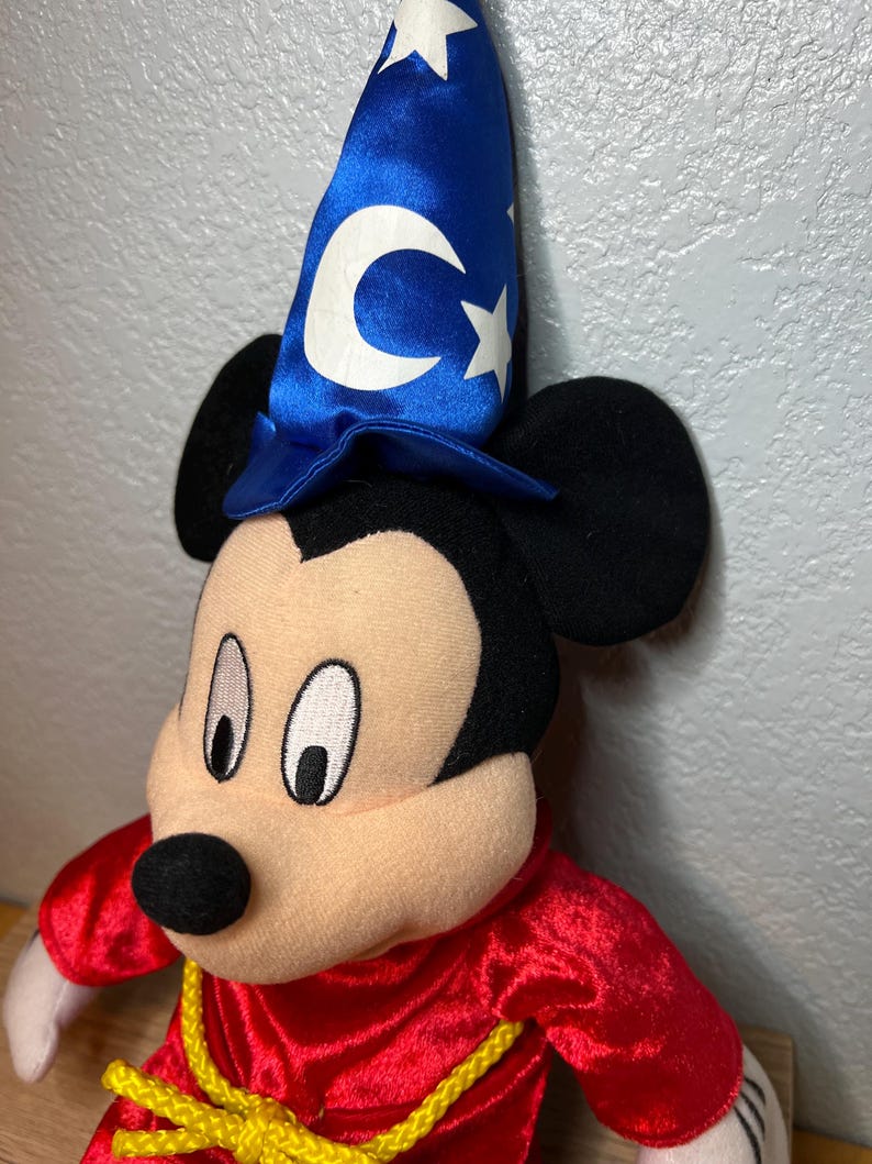 K&ouml;nnte beinhalten: Ein Mickey Mouse-Pl&uuml;schtier, das als Zauberer verkleidet ist. Das Spielzeug hat einen blauen Hut mit wei&szlig;en Sternen und einem Mond, eine rote Robe und einen gelben Seilg&uuml;rtel. Das Gesicht ist beige mit schwarzen Augen und einer schwarzen Nase.