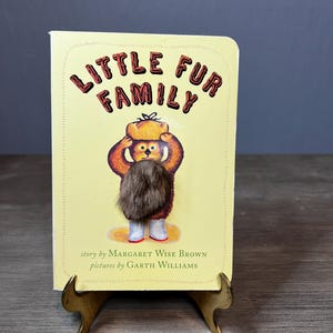May include: A children's book titled "Little Fur Family" with a yellow cover and a brown bear illustration. The book is propped up on a small, gold-colored stand. The book's cover features the title in a whimsical font.