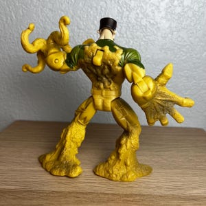 Puede incluir: Figura de acci&oacute;n amarilla con un dise&ntilde;o de camuflaje verde y amarillo en la parte superior del cuerpo. La figura tiene una gorra negra y una apariencia texturizada y derretida en los brazos y las piernas. La figura posa con un brazo extendido.