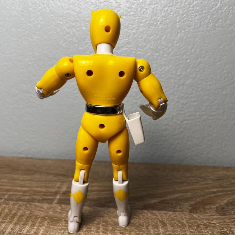Op de afbeelding: Een gele actiefiguur met witte accenten, waaronder laarzen en een holster. De figuur heeft een zwarte riem en poseert met uitgestrekte armen. De figuur is gemaakt van plastic en heeft een vintage esthetiek.