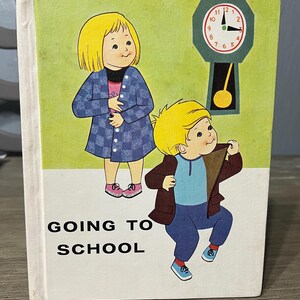 May include: Vintage children's book cover with illustrations of two children and a clock. The cover features the text "GOING TO SCHOOL" in bold, black letters. The illustrations depict a girl in a blue checkered coat and a boy in a brown jacket.
