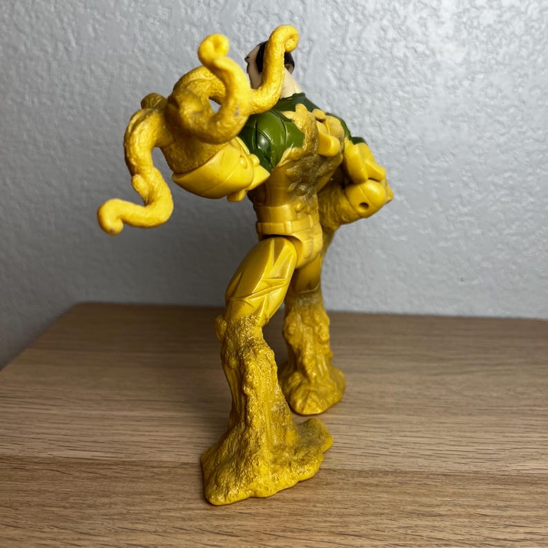 Puede incluir: Una figura de acci&oacute;n amarilla con una constituci&oacute;n musculosa y un torso verde y amarillo. La figura tiene ap&eacute;ndices amarillos en forma de tent&aacute;culos y piernas texturizadas con aspecto rocoso. La figura est&aacute; posada sobre una superficie de madera.