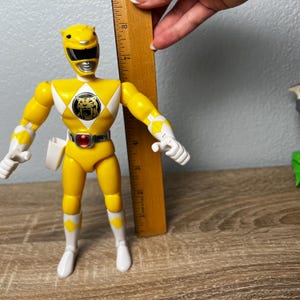 Op de afbeelding: Een gele Power Ranger actiefiguur, ongeveer 18 cm hoog, met witte en zwarte accenten. De figuur heeft een tijgerembleem op de borst en een zilveren riem met een rode edelsteen. De figuur poseert met uitgestrekte armen.