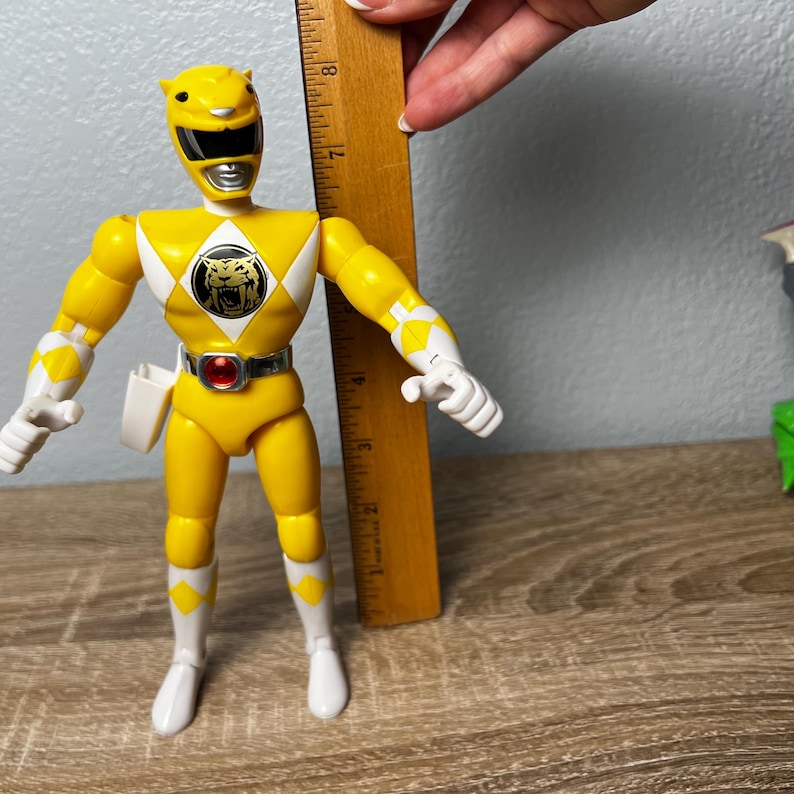 Op de afbeelding: Een gele Power Ranger actiefiguur, ongeveer 18 cm hoog, met witte en zwarte accenten. De figuur heeft een tijgerembleem op de borst en een zilveren riem met een rode edelsteen. De figuur poseert met uitgestrekte armen.