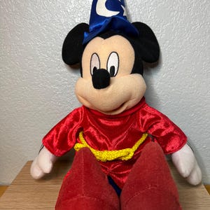K&ouml;nnte beinhalten: Ein Pl&uuml;schtier von Mickey Mouse, der als Zauberer verkleidet ist. Das Spielzeug hat einen blauen Zaubererhut mit einem wei&szlig;en Halbmond und Sternen, eine rote Robe mit einem gelben Seilg&uuml;rtel und rote Schuhe. Das Spielzeug sitzt auf einer Holzoberfl&auml;che.