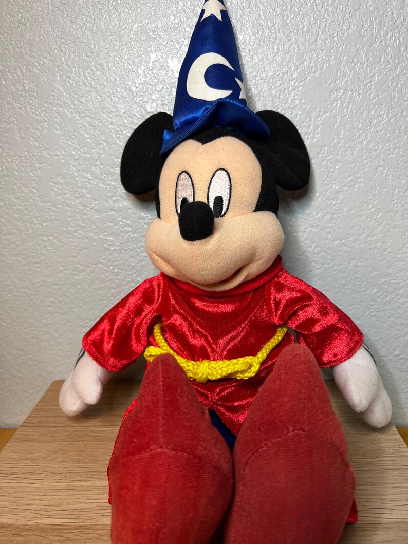 K&ouml;nnte beinhalten: Ein Pl&uuml;schtier von Mickey Mouse, der als Zauberer verkleidet ist. Das Spielzeug hat einen blauen Zaubererhut mit einem wei&szlig;en Halbmond und Sternen, eine rote Robe mit einem gelben Seilg&uuml;rtel und rote Schuhe. Das Spielzeug sitzt auf einer Holzoberfl&auml;che.