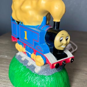 May include: A blue, red, and yellow Thomas the Tank Engine night light. The train has the number 1 on its side and a smiling face. The night light sits on a green base with a gray track.