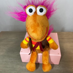 May include: A plush toy character with a bright orange body, large white eyes with black pupils, and vibrant pink hair. The toy wears a brown vest and has yellow and red striped arms. It is sitting on a pink box.