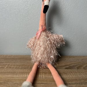 May include: A plush flamingo toy in shades of pink, with a gold crown, black beak, and silver feet. The flamingo has a fluffy, fringed chest and a pink bow. The toy is sitting on a wooden surface.