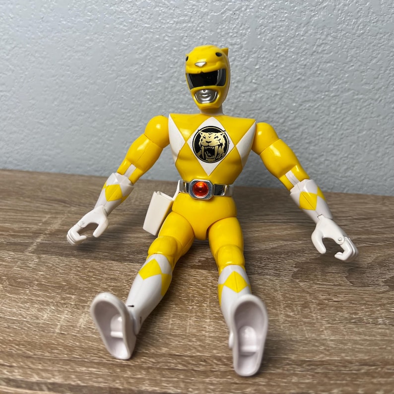 Op de afbeelding: Een gele Power Ranger actiefiguur, met witte accenten op de armen en benen. De figuur heeft een zwart vizier en een tijgerembleem op de borst. Hij zit met uitgestrekte armen.