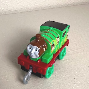 May include: A green and brown toy train with a smiling face, red accents, and green wheels. The train features a jungle-themed design with monkey illustrations. A silver hook is attached to the front of the train.