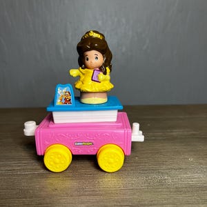 May include: A Little People toy train featuring a Belle figurine in a yellow dress, with a book in hand. The train car is pink with yellow wheels and the text "Little People". The train car is approximately 4 inches long.