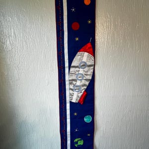 May include: A navy blue fabric growth chart with a rocket ship, planets, and stars design. The chart has a white stripe with measurement markings in inches. The rocket is silver with red accents. The planets are orange, green, and red.