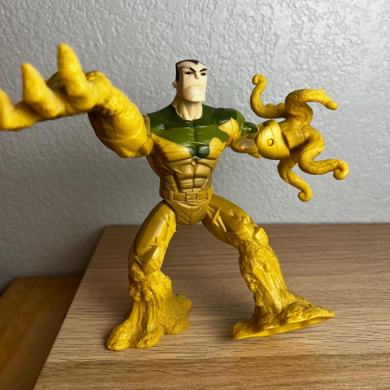 Puede incluir: Una figura de acci&oacute;n amarilla de un personaje musculoso con un torso verde y beige y una expresi&oacute;n severa. La figura tiene una apariencia texturizada y rocosa en la parte inferior del cuerpo y los brazos, y posa con un brazo extendido y el otro sosteniendo un ap&eacute;ndice con tent&aacute;culos.