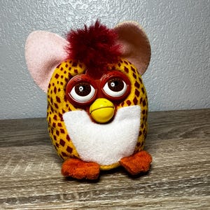 May include: A plush toy featuring a yellow body with a brown diamond pattern, a white chest patch, and orange feet. It has large brown eyes, a yellow beak, and a tuft of red hair on top. The toy includes two large, light pink felt ears.