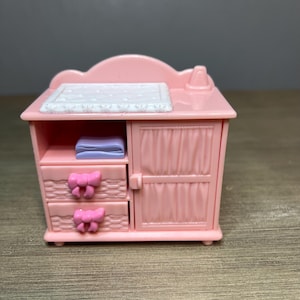 May include: A pink miniature nursery dresser with two drawers adorned with pink bow accents. The dresser has a cabinet door with vertical paneling and a white changing pad on top. A purple blanket is visible inside.