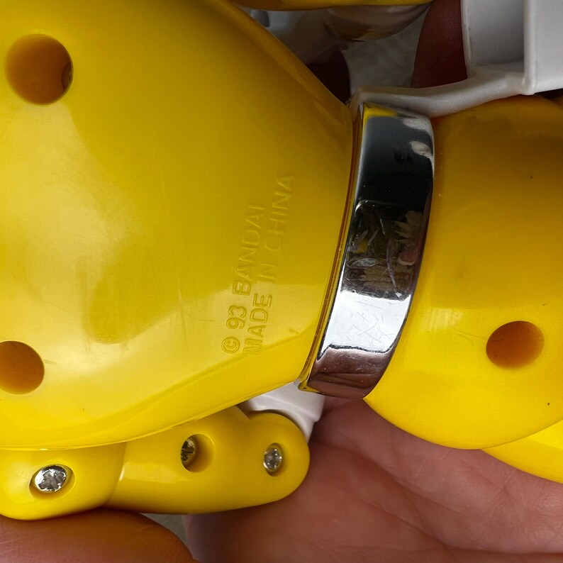 Op de afbeelding: Close-up van een geel speelgoed met een zilveren band. De tekst "&copy; 93 BANDAI MADE IN CHINA" is gedrukt op het gele plastic. Het speelgoed heeft zichtbare schroeven en een wit onderdeel.