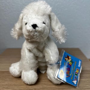 May include: A white plush toy poodle with black eyes and nose. The toy has a fluffy head and ears, and is sitting upright. A blue tag with the words "Webkinz" and "Lil' Kinz" is attached.