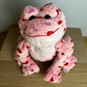 May include: A plush pink frog with red and purple spots. The frog has black eyes and a red stitched smile. The toy is sitting on a light brown wooden surface.
