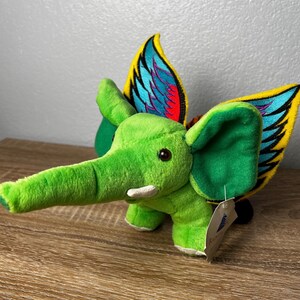 May include: A bright green plush elephant toy featuring large, soft ears and prominent, colorful wings attached to its back. The wings display vibrant yellow, blue, and red patterns. A small white tag is attached to the side of the elephant's body.