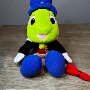 May include: A plush green cricket toy wearing a blue top hat, black jacket, and white gloves. The cricket has large black eyes, a pink mouth, and blue feet. A red bow tie is visible.
