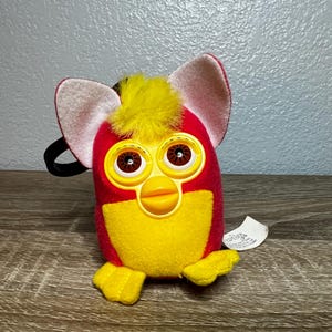 May include: A plush toy featuring a red body with a yellow belly, yellow feet, and a yellow tuft of hair on top. The toy has large, round, brown eyes surrounded by yellow plastic frames, a yellow beak, and two large, light pink felt ears.