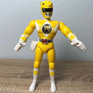 Op de afbeelding: Een gele actiefiguur van een Power Ranger met een tijgerembleem op de borst. De figuur heeft een gele helm, witte handschoenen en witte laarzen. Een zilveren riem met een rode edelsteen zit om de taille. De figuur is ongeveer 13 cm hoog.