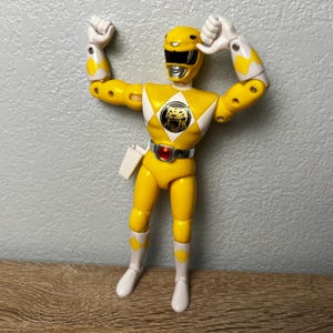 Op de afbeelding: Een gele actiefiguur van een Power Ranger met een tijgerembleem op de borst. De figuur draagt een gele helm, witte laarzen en een gereedschapsriem. De armen zijn geheven in een feestelijke pose.