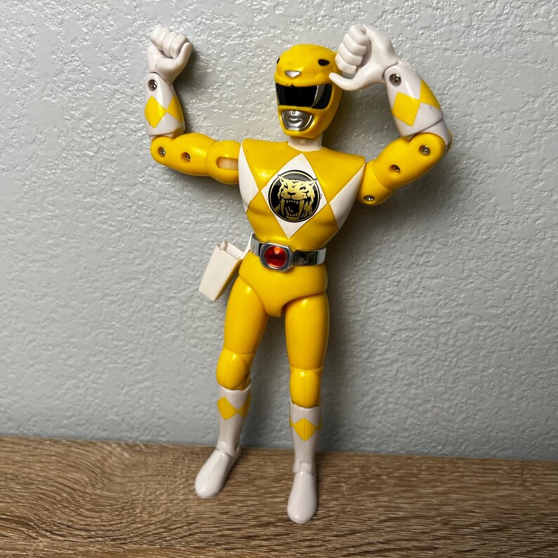 Op de afbeelding: Een gele actiefiguur van een Power Ranger met een tijgerembleem op de borst. De figuur draagt een gele helm, witte laarzen en een gereedschapsriem. De armen zijn geheven in een feestelijke pose.