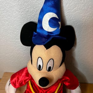 K&ouml;nnte beinhalten: Ein Pl&uuml;schtier von Mickey Mouse als Zauberer verkleidet. Mickey tr&auml;gt einen blauen Zaubererhut mit einem wei&szlig;en Halbmond und Stern, eine rote Robe und einen gelben Seilg&uuml;rtel. Das Spielzeug hat schwarze Ohren, ein beiges Gesicht und wei&szlig;e Handschuhe.