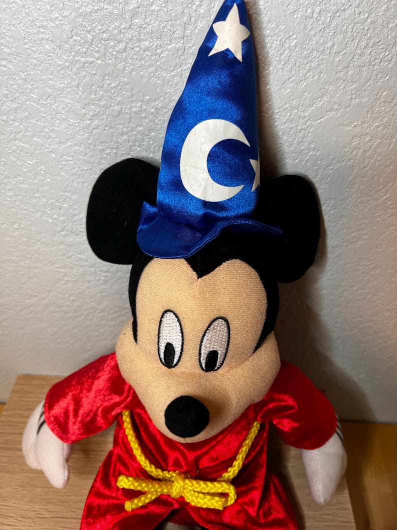 K&ouml;nnte beinhalten: Ein Pl&uuml;schtier von Mickey Mouse als Zauberer verkleidet. Mickey tr&auml;gt einen blauen Zaubererhut mit einem wei&szlig;en Halbmond und Stern, eine rote Robe und einen gelben Seilg&uuml;rtel. Das Spielzeug hat schwarze Ohren, ein beiges Gesicht und wei&szlig;e Handschuhe.