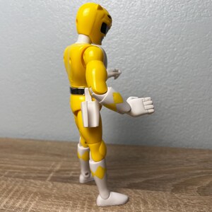 Op de afbeelding: Een gele actiefiguur van een Power Ranger, met witte en gele accenten. De figuur draagt een helm en een riem. De figuur staat op een houten oppervlak.