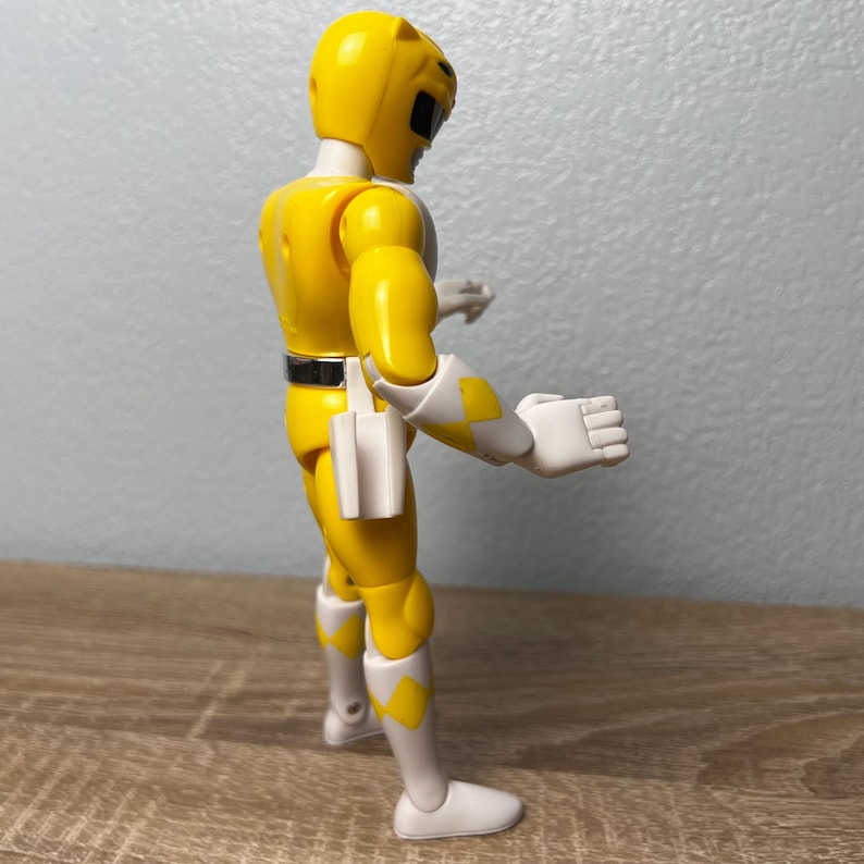 Op de afbeelding: Een gele actiefiguur van een Power Ranger, met witte en gele accenten. De figuur draagt een helm en een riem. De figuur staat op een houten oppervlak.