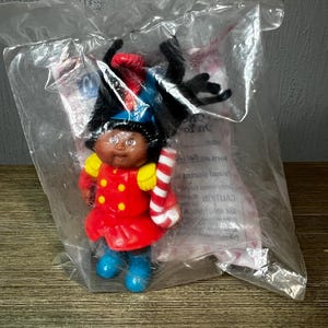 May include: A Cabbage Patch Kids doll in a clear plastic bag. The doll is dressed in a red jacket with gold buttons, blue boots, and a black hat with a red plume. It holds a candy cane.