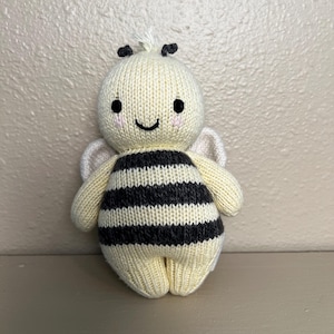 May include: A handmade, knitted bee doll with a cream-colored body and black and gray striped abdomen. The bee has small black eyes, rosy cheeks, and a smiling mouth. It has small wings and antennae.
