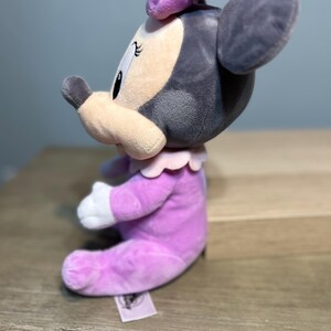 Puede incluir: Mu&ntilde;eca de peluche de Minnie Mouse sentada. La mu&ntilde;eca lleva un vestido y un lazo de color lavanda, con un cuello rosa claro y guantes blancos. La cara es de color rosa claro con ojos negros y una nariz negra. Las orejas son grises.