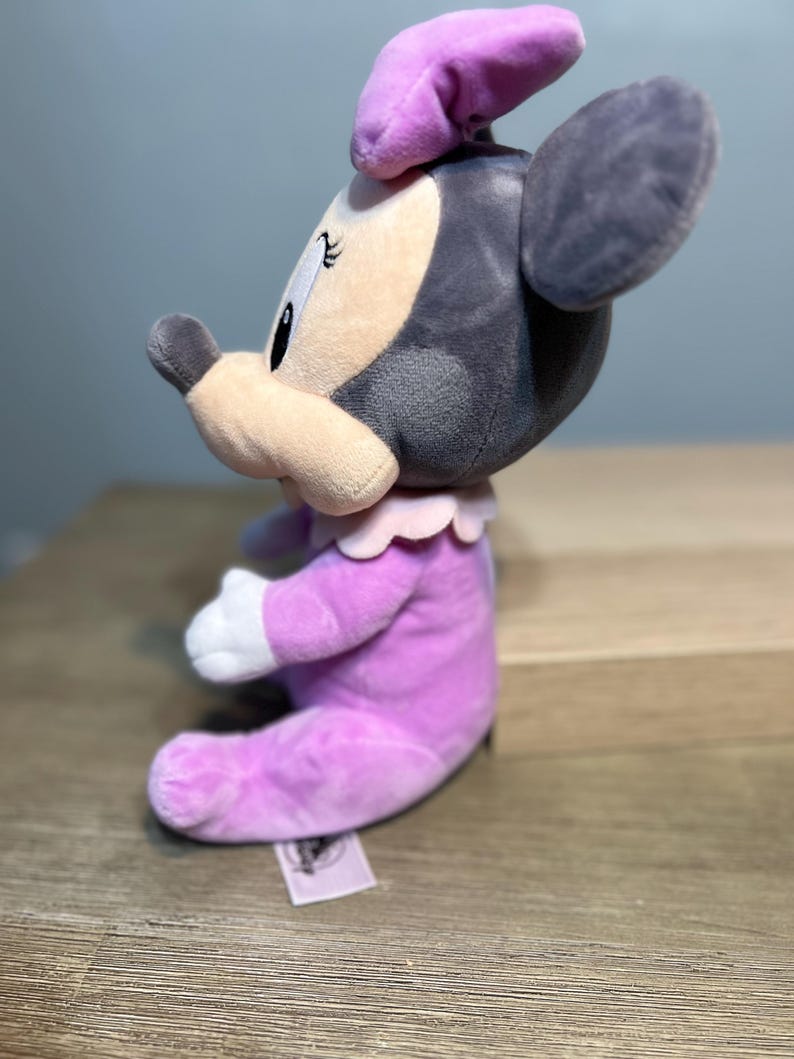 Puede incluir: Mu&ntilde;eca de peluche de Minnie Mouse sentada. La mu&ntilde;eca lleva un vestido y un lazo de color lavanda, con un cuello rosa claro y guantes blancos. La cara es de color rosa claro con ojos negros y una nariz negra. Las orejas son grises.