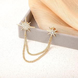 May include: A gold-toned brooch featuring two starburst designs connected by delicate chains. The starbursts are embellished with clear crystals, adding sparkle. The brooch is presented on a neutral surface with a soft, golden fabric in the background.