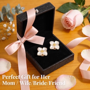May include: A pair of white flower-shaped stud earrings with yellow centres, presented in a black textured jewellery box with a light pink satin ribbon tied around the lid. The text "Perfect Gift for Her Mom / Wife / Bride / Friend" is visible.