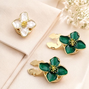 Op de afbeelding: Drie bloemvormige broches. Eén is wit met een gouden rand en midden, de andere twee zijn smaragdgroen met gouden randen en middens. De broches liggen op een crèmekleurig oppervlak.