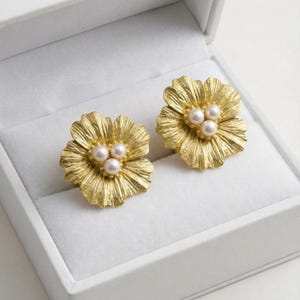 May include: A pair of gold-colored flower-shaped earrings, each with three small, round pearls at the center. The earrings are displayed inside a white jewelry box, showcasing their intricate petal design and pearl accents.
