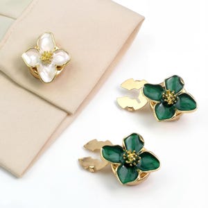 May include: Floral-shaped cufflinks. One cufflink is white with a gold border and center. Two cufflinks are green with gold borders and centers. The cufflinks are on a white background.