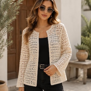 May include: A cream-colored, open-front crochet cardigan featuring a repeating square grid pattern, long sleeves, and a round neckline. The garment is worn over a solid black top with a black belt, creating a contrast between the light texture and dark base.