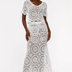 May include: A white two-piece crochet set consisting of a short-sleeved top with a V-neckline and tie-detail sleeves, paired with a matching floor-length maxi skirt. Both pieces feature a repeating circular mandala-style pattern throughout.