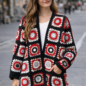 Op de afbeelding: Een zwarte, openvallende cardigan met een kleurrijk granny square patroon. De vierkanten hebben rode, witte en grijze accenten. De cardigan heeft lange mouwen en zakken. Het kledingstuk wordt gedragen over een witte top en een blauwe jeans.