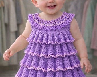 May include: A smiling baby wearing a handmade, tiered purple crochet dress and matching headband. The dress features a scalloped neckline and edges, with a lighter purple trim. The background shows a closet with hanging clothes.