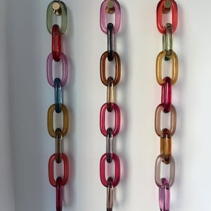 以下が含まれることがあります： 半透明のマルチカラーアクリルリンクで作られた3本の垂直な装飾チェーン。各チェーンは小さな円形のゴールドトーンの壁掛けフックから吊り下げられています。リンクは赤、ピンク、青、緑、琥珀色など、さまざまな鮮やかな色合いを特徴としています。