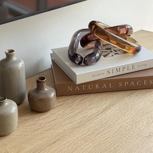 May include: A decorative glass chain in amber and purple tones sits atop books titled "modern SIMPLE living" and "NATURAL SPACES." Three ceramic vases in neutral colours are arranged nearby, creating a minimalist aesthetic.
